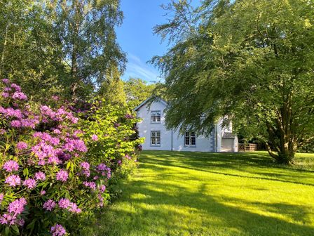 A white house with multiple windows surrounded by trees and purple flowering bushes with a green lawn at The Vicarage near Rosthwaite