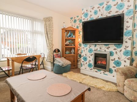 A living room with a wall-mounted TV above a fireplace a wooden cabinet with decorative items a small dining table with chairs and a beige sofa at Helredale Retreat Whitby
