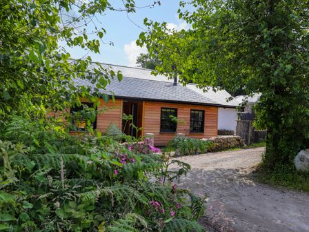 A wooden house with black framed windows next to a gravel driveway surrounded by greenery and flowers at The Dairy in Trenear near Helston