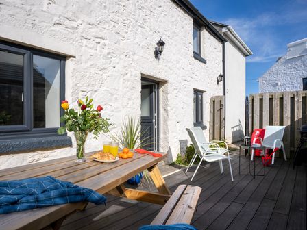 An outdoor patio with a wooden picnic table holding flowers juice croissants and oranges and white chairs with a small table at Rose Cottage in St Ives
