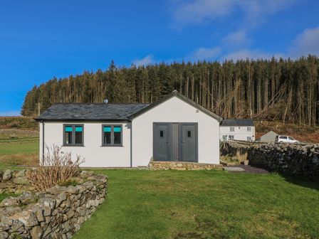 A single story house with gray doors and windows surrounded by stone walls and grass with a forest in the background at The Sail Loft in Nefyn