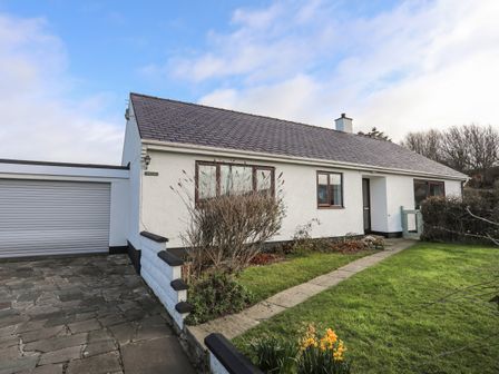 A white house with a stone driveway and a garden with green grass and flowers at Bwthyn Y Bae in Church Bay near Llanfaethlu