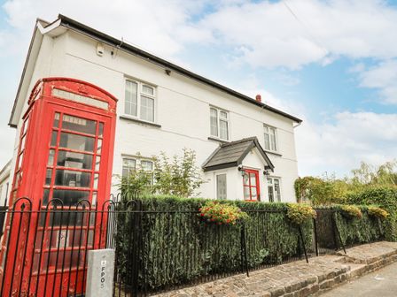 A white house with multiple windows and a red door next to a red telephone booth and black metal fence with plants