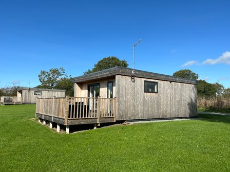 A wooden cabin with a deck and railing on a grassy area with another cabin in the background at The Wren in Wem