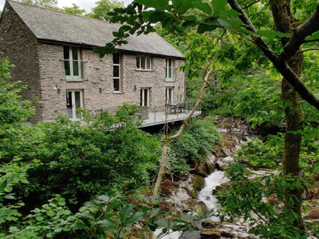 A stone house with multiple windows and doors next to a stream surrounded by trees at The Old Water Mill in Kendal
