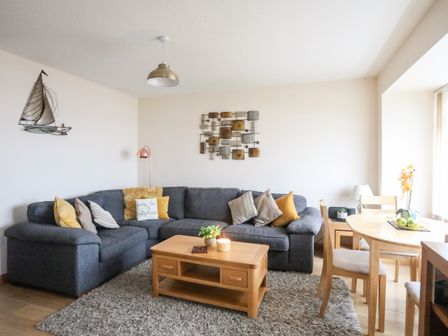A living room with a dark sectional sofa and wooden coffee and dining tables at Sandy Toes in Rhosneigr