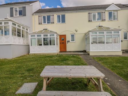 A wooden picnic table on grass in front of a building with white conservatories and a wooden door at Grooms Cottage in St Florence
