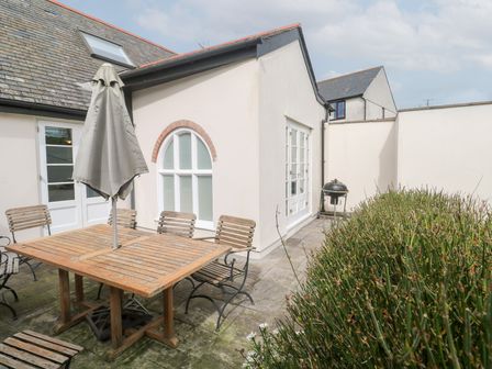 An outdoor patio with wooden table and chairs and a closed umbrella next to a white house with arched and glass doors and a barbecue grill at No 3 The Hinges in Crantock