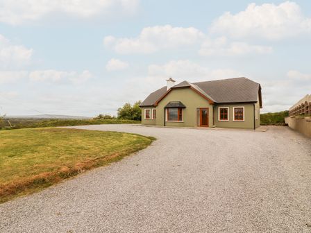 A single-story house with green exterior walls and a gray roof on a gravel driveway with a grassy area beside it at Daisys Cottage in Knockreagh near Listowel County Kerry