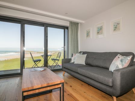 A living room with a gray sofa wooden coffee table and balcony with two chairs overlooking the beach at Fistral Watch in Newquay