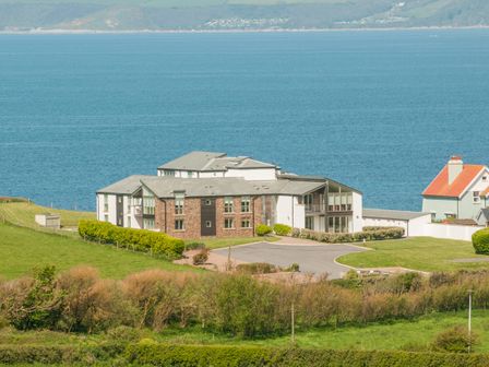 A building near the ocean with grassy areas and a house with an orange roof in the background at 10 Oceans Edge in Thurlestone
