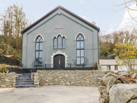 A stone building with arched windows and a gated entrance surrounded by a stone wall at Greystones Chapel in Cwm-y-Glo near Llanrug