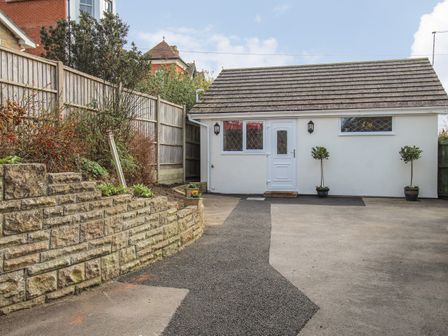 A small white building with a shingled roof and two small trees in pots on either side of the door at Roundham Cottage in West Bay