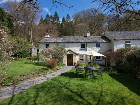 A stone path leading to a cottage with garden furniture and an umbrella on a lawn at Fir Tree Cottage in Grasmere