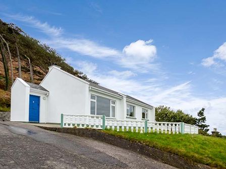 A white house with a blue door on a hillside with green grass and trees at Rossbeigh Beach Cottage No 4 Rossbeigh near Glenbeigh County Kerry