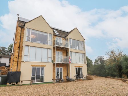 A multi-story building with large windows and balconies with outdoor chairs on a gravel area surrounded by trees at Cotswold View in Bourton-On-The-Water