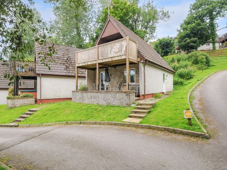 A triangular roof house with balcony and patio chairs on grass near a curved road at Dart Lodge in Callington