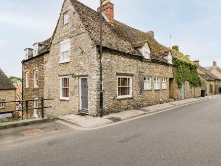 A stone cottage with multiple windows and a slate roof on a street at Corner Cottage in Malmesbury