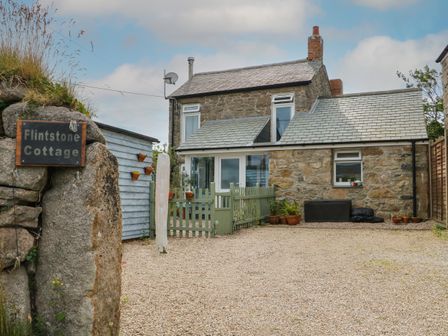 A stone cottage with a gravel driveway green picket fence and potted plants at Flintstone Cottage in Pendeen