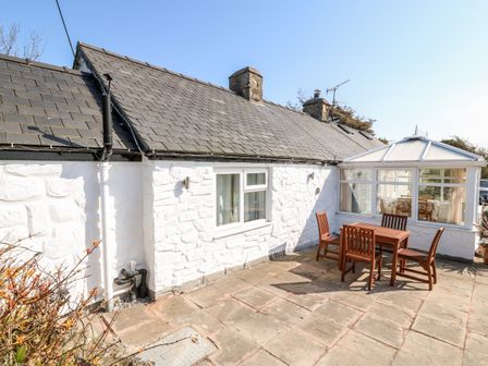 A white stone exterior cottage with a slate roof and a wooden outdoor table with chairs on a stone patio at Y Big in Morfa Nefyn