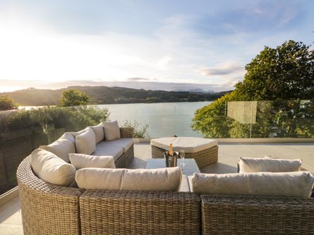 An outdoor patio with wicker sectional sofa and ottoman overlooking a lake with hills and trees at Kessock in Bowness-On-Windermere