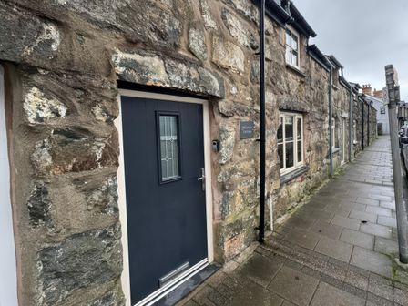 A stone cottage with a dark blue door and windows along a paved sidewalk at Weavers Cottage Tremadog