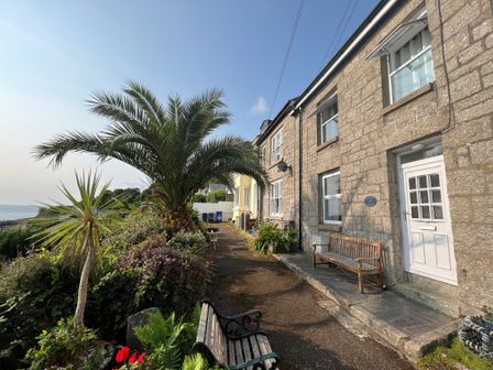 A stone house with white doors and windows beside a garden with palm trees and benches at Lookout Cottage in Newlyn