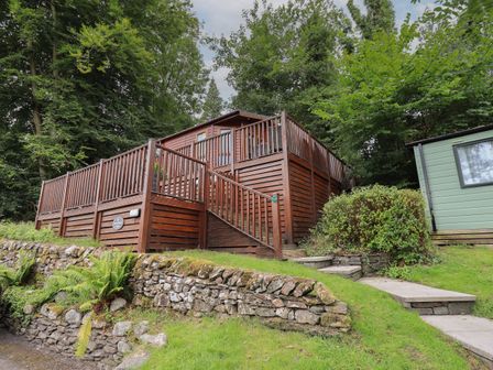 A wooden lodge with a deck and stairs surrounded by greenery and stone walls at Acorn Bank Lodge L 55 Fallbarrow Park near Bowness-On-Windermere