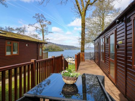 A deck with wooden railings and a table with a flower basket between two wooden lodges by a lake at Shoreside Lodge L8 Fallbarrow Park near Bowness on Windermere