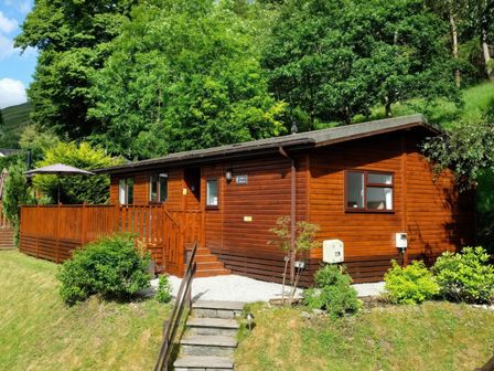 A wooden cabin with a deck and umbrella surrounded by trees and bushes at Walkers Retreat Lodge Fellside 5 Limefitt Park near Windermere