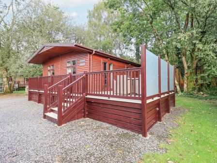 A wooden lodge with a raised deck and stairs surrounded by trees and gravel at Tanglewood Lodge Thirlmere 15 White Cross Bay near Windermere