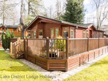 A wooden lodge with a fenced deck surrounded by grass and trees at Broad Larch Lodge Glade 16 Fallbarrow Park near Bowness on Windermere