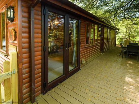 A wooden cabin exterior with glass double doors and outdoor dining table on a deck surrounded by trees at Lakeshaven Skiptory Howe 16 White Cross Bay near Windermere
