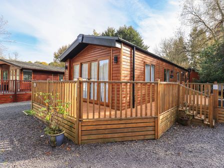 An exterior view of a wooden lodge with a wooden deck and railing surrounded by gravel at Glenridding Lodge Grasmere 2 White Cross Bay near Windermere