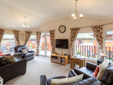 A living room with leather sofas a wall clock a TV on a stand and large windows with curtains at Hideaway Lodge Kirkstone 14 Limefitt Park near Windermere