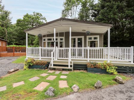 A front view of a small cabin with a covered porch and white railing surrounded by grass and trees at Pinehaven Lodge in Grasmere 6 White Cross Bay near Windermere