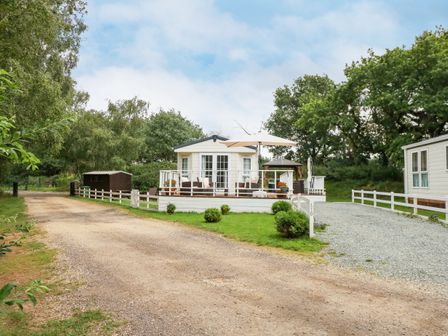A small light-colored house with a deck and outdoor furniture next to a gravel driveway with trees in the background at Lakeside View in Pentney near King's Lynn