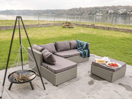 An outdoor patio with a grey sectional sofa with cushions and a blanket a matching ottoman with a plate of strawberries and books and a tripod fire pit with wood around Bwthyn Siliwen Old Bath House in Bangor