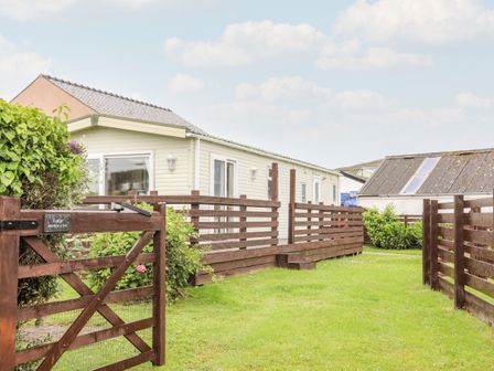 A fenced garden with a wooden gate and a single-story light-colored lodge with a deck at Bwthyn Y Bae Lodge in Church Bay near Llanfaethlu