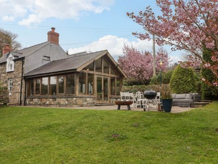 A stone and wood house with a glass extension and patio furniture surrounded by green lawn and flowering trees at Sea View in Newport Pembrokeshire