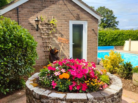 A round stone planter with colorful flowers near a brick building with a white door and a carved bear holding a welcome sign at The Pool House in Crediton