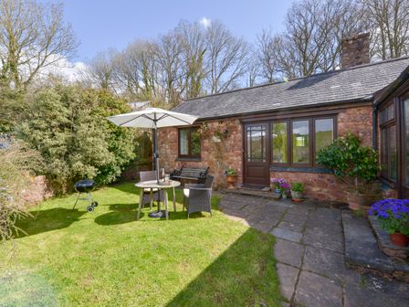 A garden with a stone house a table and umbrella chairs a barbecue and flower pots at Little Barton in Stogumber near Watchet