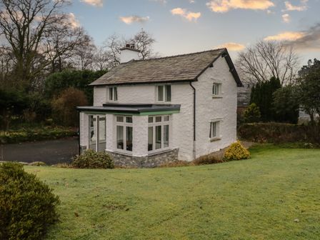 A white stone cottage with a slate roof and a glass porch surrounded by grass and trees at Green Stile Cottage in Bowness-On-Windermere