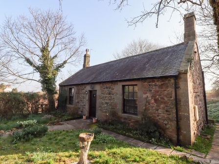 A stone cottage with two windows and a door surrounded by a garden and trees at Rose Cottage Akeld near Wooler