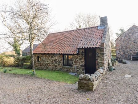 A stone cottage with a red tiled roof surrounded by a gravel driveway and a small lawn with a tree at Tile Rouge in Wooler