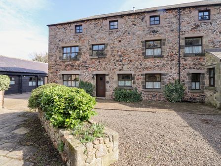 Exterior view of a stone building with multiple windows and a wooden door with bushes in a stone planter at Ivy Cottage in Akeld near Wooler