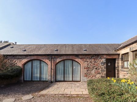 A stone cottage with two arched windows and a wooden door surrounded by bushes and daffodils at Pansy Plot in Akeld near Wooler