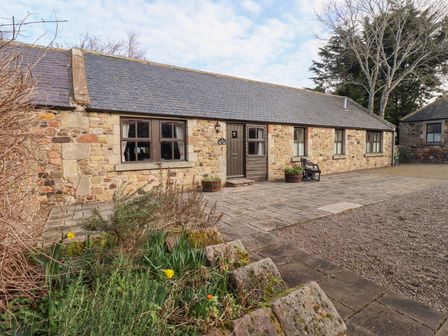 Exterior view of a single story stone cottage with multiple windows and a wooden door surrounded by paved stones and plants at Heatherview Akeld near Wooler
