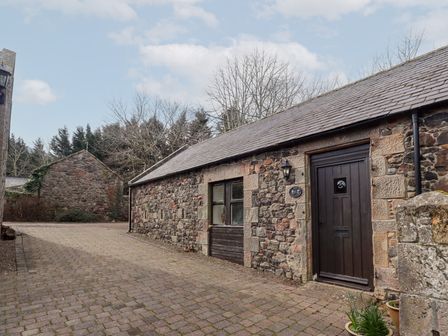 A stone building with a dark wooden door and window beside a paved courtyard at The Columbine in Akeld near Wooler