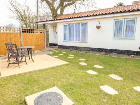 An outdoor garden area with a paved patio table and two chairs lawn with stepping stones and a single-story building with windows at Jimmy's Place in Pulham Market